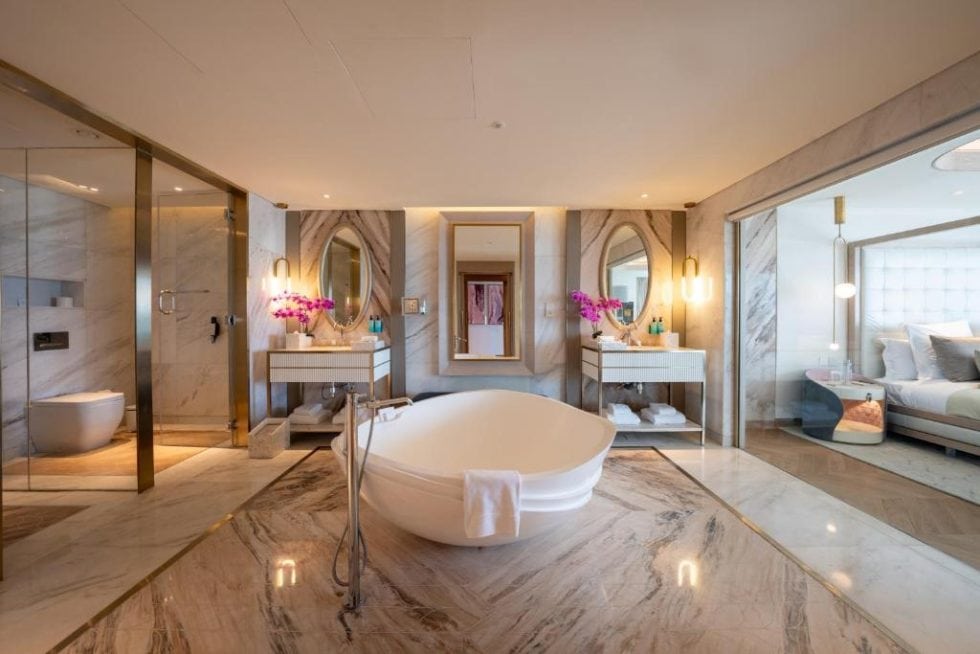 Luxueuse salle de bain avec baignoire îlot, doubles lavabos, miroir rond, décoration élégante, touches de fleurs violettes, espace de détente et vue sur la chambre et la salle de bain.
