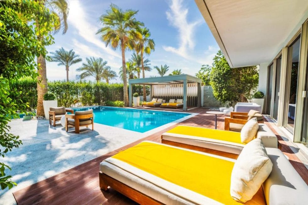 Luxueuse terrasse avec piscine privée, chaises longues et espace détente sous un ciel bleu ensoleillé, entourée de palmiers et de végétation luxuriante.